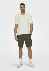 Camisa polo de manga corta color beige claro con cuello, combinada con pantalones cortos verdes oscuros. El modelo lleva zapatillas blancas con detalles en verde.