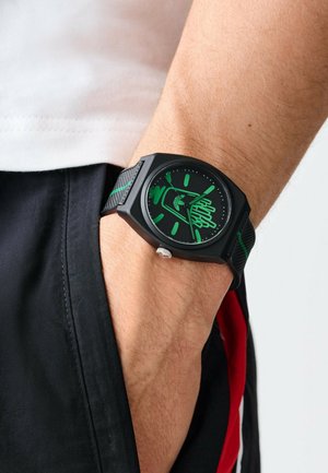 Montre noire au design géométrique, avec des accents verts et un bracelet texturé. Le cadran de la montre présente de grands index d'heure et des aiguilles verts.
