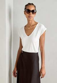 Biała koszulka z dekoltem w serek z podwiniętymi rękawami, w połączeniu z czarną spódnicą w stylu wrap do kolan, z bocznym detalem na guziki. Modelka nosi ciemne okulary przeciwsłoneczne.