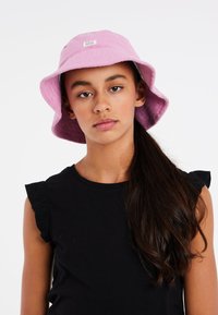 Rosa bucket hat lavet af frotté med et lille logo-tag, der har en bred skygge og blød tekstur, parret med en sort top med flæser på ærmerne.