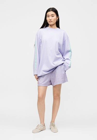 Νεαρή γυναίκα στέκεται φορώντας ένα ανοιχτό μωβ φαρδύ πουκάμισο με μακριά μανίκια, μοβ καρό σορτς, μπεζ αθλητικά παπούτσια, με ουδέτερη έκφραση.