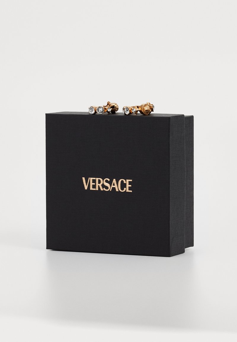Goldfarbene Ohrringe mit klaren Edelsteinen auf einer strukturierten schwarzen Box mit goldfarbener "VERSACE"-Markierung.
