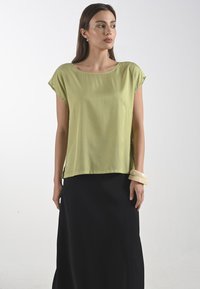 Limetkově zelená blůza s krátkým rukávem a širokým výstřihem, spárovaná s černou dlouhou sukní. Hladká textura, volný střih a lehký materiál.