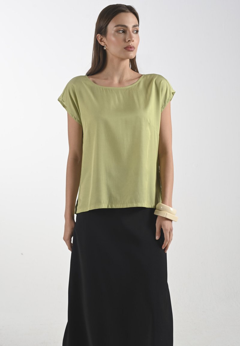 Limetkově zelená blůza s krátkým rukávem a širokým výstřihem, spárovaná s černou dlouhou sukní. Hladká textura, volný střih a lehký materiál.