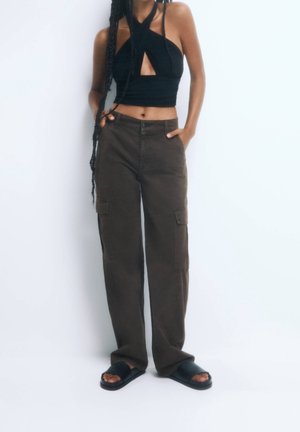 Persoon met een zwarte halter crop top, donkerbruine cargobroek en zwarte slidesandalen, staand met handen in de zakken tegen een witte achtergrond.