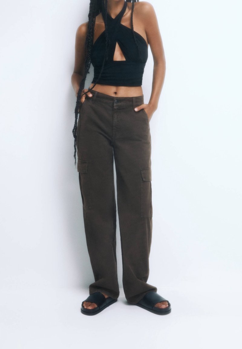 Personne portant un crop top noir à col halter, un pantalon cargo marron foncé et des sandales slides noires, debout avec les mains dans les poches sur un fond blanc.