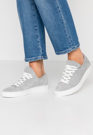 Person iført grå ruskindssneakers med hvide snørebånd og såler, kombineret med cropped blå denimjeans mod en ensfarvet baggrund.