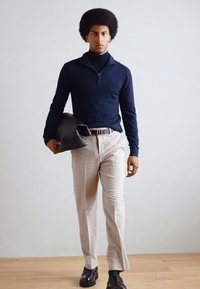 Suéter navy con cuello alto, pantalones beige claro a medida y zapatos de cuero negros. El modelo sostiene una chaqueta gris oscuro. Texturas suaves.
