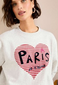 Sivý sveter s grafickým prvkom červeného pruhovaného srdca s nápisom "PARÍŽ" v čiernej farbe a "je t'aime" v modrej farbe. Okrúhly výstrih a mäkká textúra.