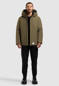 Olive green hooded jacket with a smooth texture and front zipper. Paired with black cargo pants and chunky black boots.