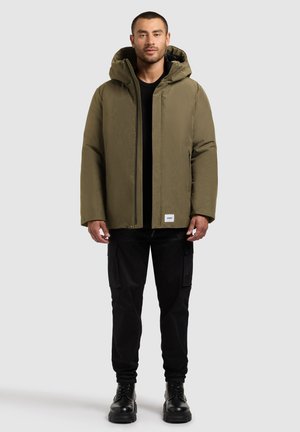 Olijfgroene hoodie-jack met een gladde textuur en een ritssluiting aan de voorkant. Gepaard met zwarte cargobroek en stevige zwarte laarzen.