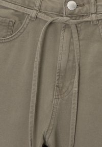 Gros plan sur la taille d'un pantalon kaki avec passants pour ceinture, un cordon de serrage, une poche et un bouton argenté avec le nom de la marque.