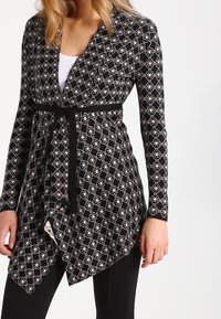 Kvinna som bär en långärmad cardigan med svart-vitt mönster och ett svart bälte över en vit topp och svarta byxor.