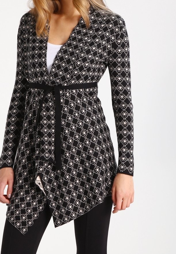 Kvinna som bär en långärmad cardigan med svart-vitt mönster och ett svart bälte över en vit topp och svarta byxor.