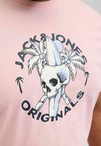 Hele roosa puuvillane T-särk, millel on graafika, mis kujutab kolju, surfilauda ja palmipuid. Tekst: "JACK & JONES ORIGINALS."