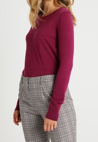 Fuksjowy sweter z długim rękawem i okrągłym dekoltem, połączony z wysokimi spodniami w kratę w kolorze czarnym, białym i szarym. Lekka dzianina.