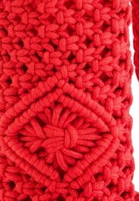 Tissu en crochet rouge présentant un motif de diamant proéminent avec des nœuds complexes et une finition texturée. Le matériau semble doux et épais.