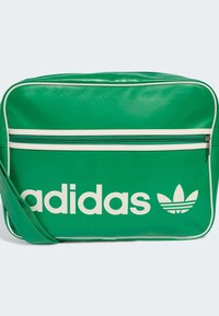 Grønn vinylveske med hvite detaljer, stort hvitt "adidas"-logo og det ikoniske trefoil-logoet. Har en frontlomme med glidelås.