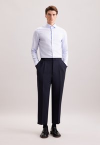 Camicia a quadri blu chiaro e bianca, abbinata a pantaloni eleganti blu scuro e scarpe nere da uomo, indossata con le mani in tasca.