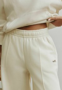 Kreemivärvi dressipluus ja sobivad dressipüksid, millel on tikitud "self." logo, elastne vöökoht ja pehme, tekstuuriga kangas.