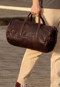 Tumši brūnas ādas ceļojumu soma ar divām izturīgām rokturām, noapaļota forma un smalka tekstūra. Redzama rāvējslēdzēja aizdares un mazas birkas detaļas.