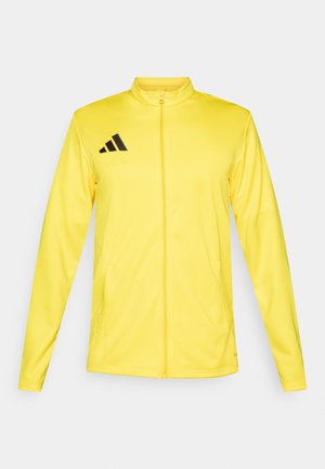 Chaqueta deportiva amarilla con cuello alto, cremallera completa y dos bolsillos laterales. Presenta un logo negro de Adidas en la parte superior izquierda.
