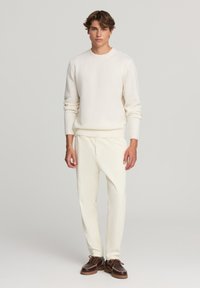 Kremowy sweter z prążkowanymi mankietami i dołem, zestawiony z teksturowanymi kremowymi spodniami oraz brązowymi butami z sznurowadłami i detalami w szyciu.