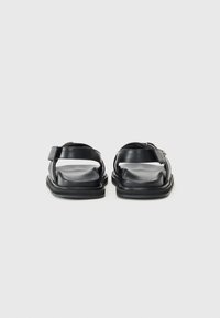 Black leather sandals featuring a flat sole, adjustable strap with buckle, and open back design. Smooth texture with minimal detailing.