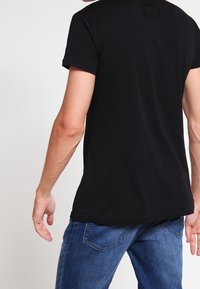 Camiseta negra de manga corta con cuello redondo, que presenta una pequeña etiqueta cuadrada en la parte posterior, combinada con jeans azules. Textura de tela suave.