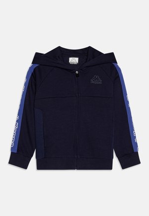 Haut à capuche zippé bleu marine en tissu doux. Doté d'une capuche, de poches et de bandes latérales bleues avec un logo. Poignets et ourlet côtelés. Texture lisse.
