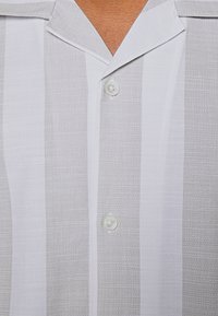 Gros plan d'une chemise à col avec des rayures verticales gris clair et blanc, avec deux boutons blancs fermés à l'avant.
