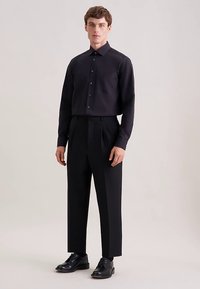 Camicia nera a maniche lunghe con colletto a punta, chiusura frontale con bottoni e tasca, abbinata a pantaloni neri a vita alta a pieghe e scarpe nere.