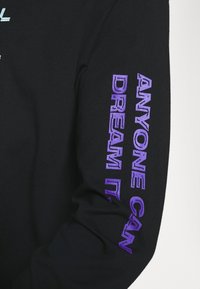 Svart långärmad tröja med lila text på ärmen som säger "VEM SOM HELST KAN DRÖMME DET." Tillverkad av mjukt tyg med en bekväm passform.