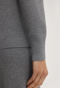 Grijze gebreide trui met geribde manchetten en onderrand, met een zachte textuur en een aansluitend ontwerp. Close-up van arm- en handplaatsing.