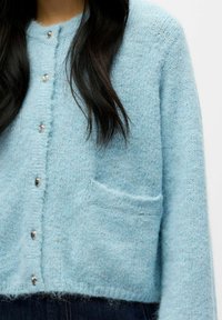 Svijetloplava pletena kardigan jakna s okruglim izrezom, bisernim gumbima i prednjim džepom, koja se odlikuje mekom, puhastom teksturom.