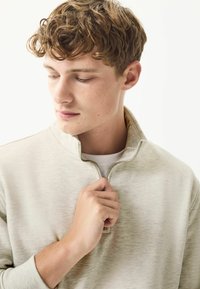 Light gray zip-up sweatshirt with a textured finish, partially zipped at the collar, worn over a white shirt, model holding the fabric.