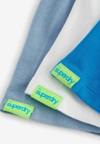 Három rövid ujjú póló kék, fehér és világoskék színben, zöld "superdry" címkékkel az alján, sima pamutszerkezettel.