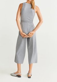 Combinaison grise à rayures avec un corsage ajusté, des jambes larges et courtes, et une taille cintrée. Portée avec des chaussures plates grises à bouts ouverts.