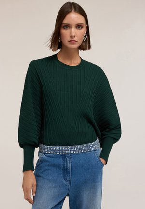 Maglione a maglia verde scuro con texture a costine, scollatura rotonda e maniche voluminose, abbinato a jeans di denim azzurri e una cintura con borchie.