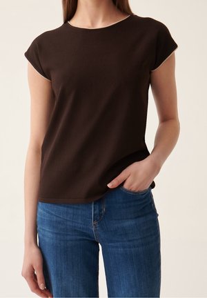 Kobieta ubrana w ciemnobrązową bluzkę z krótkim rękawem i jasnym wykończeniem, połączoną z niebieskimi dżinsami, jedną ręką w kieszeni, stojąca na jednolitym tle.