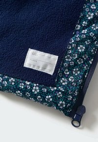 Marineblå fleece-tyg med en blomsterprydd kant; inkluderar en vit etikett för namn och telefonuppgifter, med dragkedjestängning.