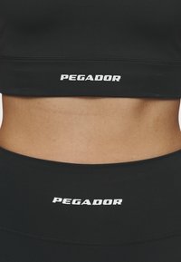 Top corto atlético negro y cinturilla que presenta el texto "PEGADOR" en blanco. Textura suave con un diseño ajustado y sin patrones visibles.