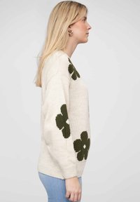 Cream sweater with green floral design on sleeves, knitted texture, relaxed fit, round neckline, and ribbed cuffs. Paired with light blue jeans.
