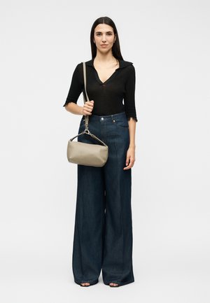 Žena s dlhými tmavými vlasmi v čiernom topu a tmavých širokých rifliach, drží béžovú kabelku na ramene pred jednoduchým svetlým pozadím.