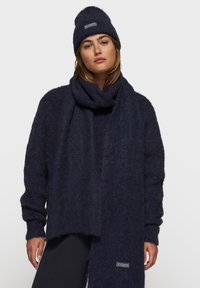 Donkerblauwe gebreide muts en sjaal set, met een zachte, fuzzy textuur en een zichtbaar merkembleem op de sjaal. Gevormd voor comfort en warmte.