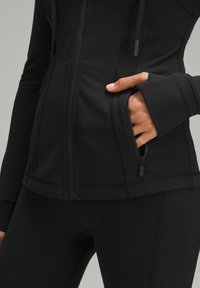 Detailní záběr ruky v kapsičce na zip přiléhavé černé sportovní bundy, která je na sobě s odpovídajícími černými legínami.