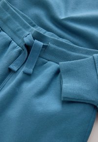 Tirkizne trenirka hlače s elastičnim pojasom i vezicom, rebrastim manšetama te mekom, glatkom teksturom. Bez vidljivih uzoraka ili dodatnih detalja.