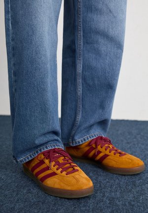 Personne portant un jean bleu et des baskets jaune moutarde avec des rayures et des lacets rouge foncé, debout sur un tapis bleu.