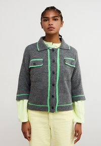 Giacca grigia lavorata a maglia con maniche corte, cuciture a contrasto verde, due tasche sul petto e bottoni neri, indossata sopra una maglietta giallo chiaro.