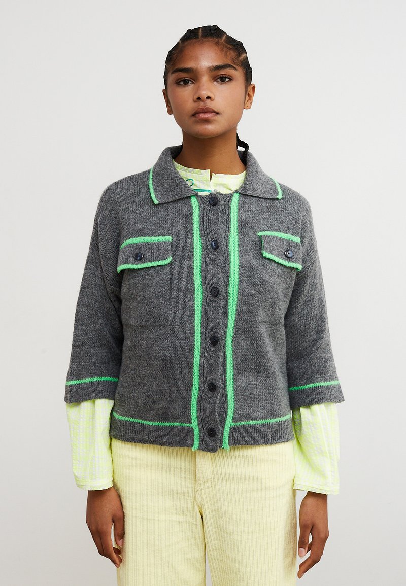 Giacca grigia lavorata a maglia con maniche corte, cuciture a contrasto verde, due tasche sul petto e bottoni neri, indossata sopra una maglietta giallo chiaro.